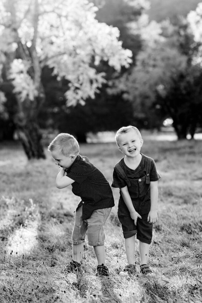 Twin boys, both with autism, stand next to each other in a grassy field, one of them turned away from the camera and another one flashing a bright grin.