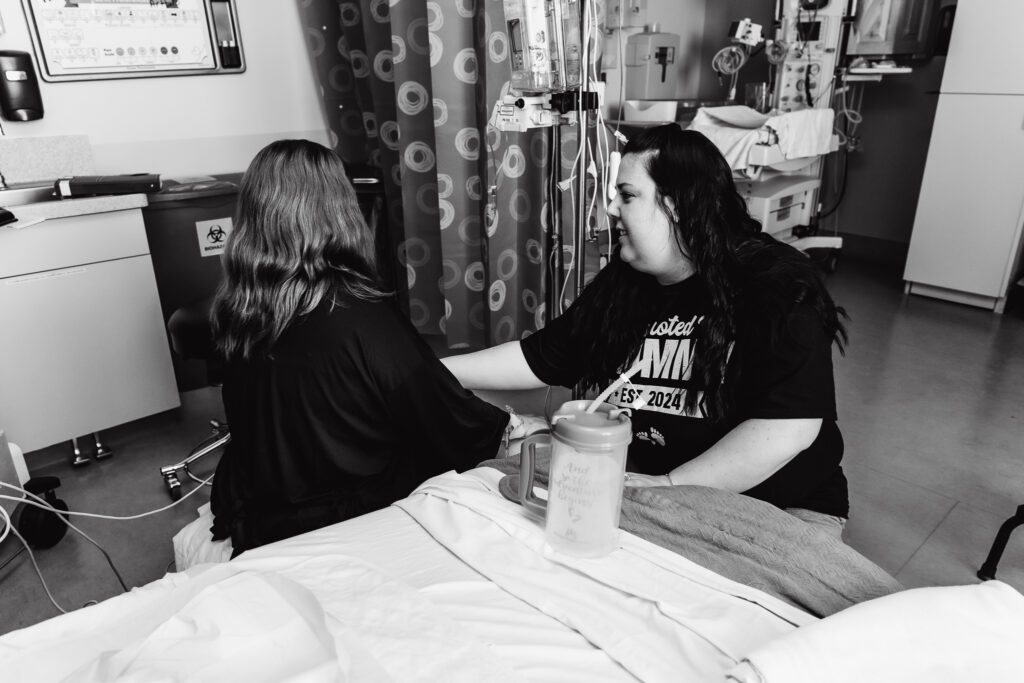 A friend holds the hand of another friend who is sitting in a hospital room 