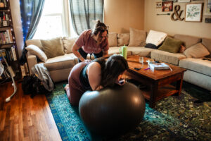 doula provides compressions on birthing mom's back as she leans over a birthing ball listening to a pregnancy podcast