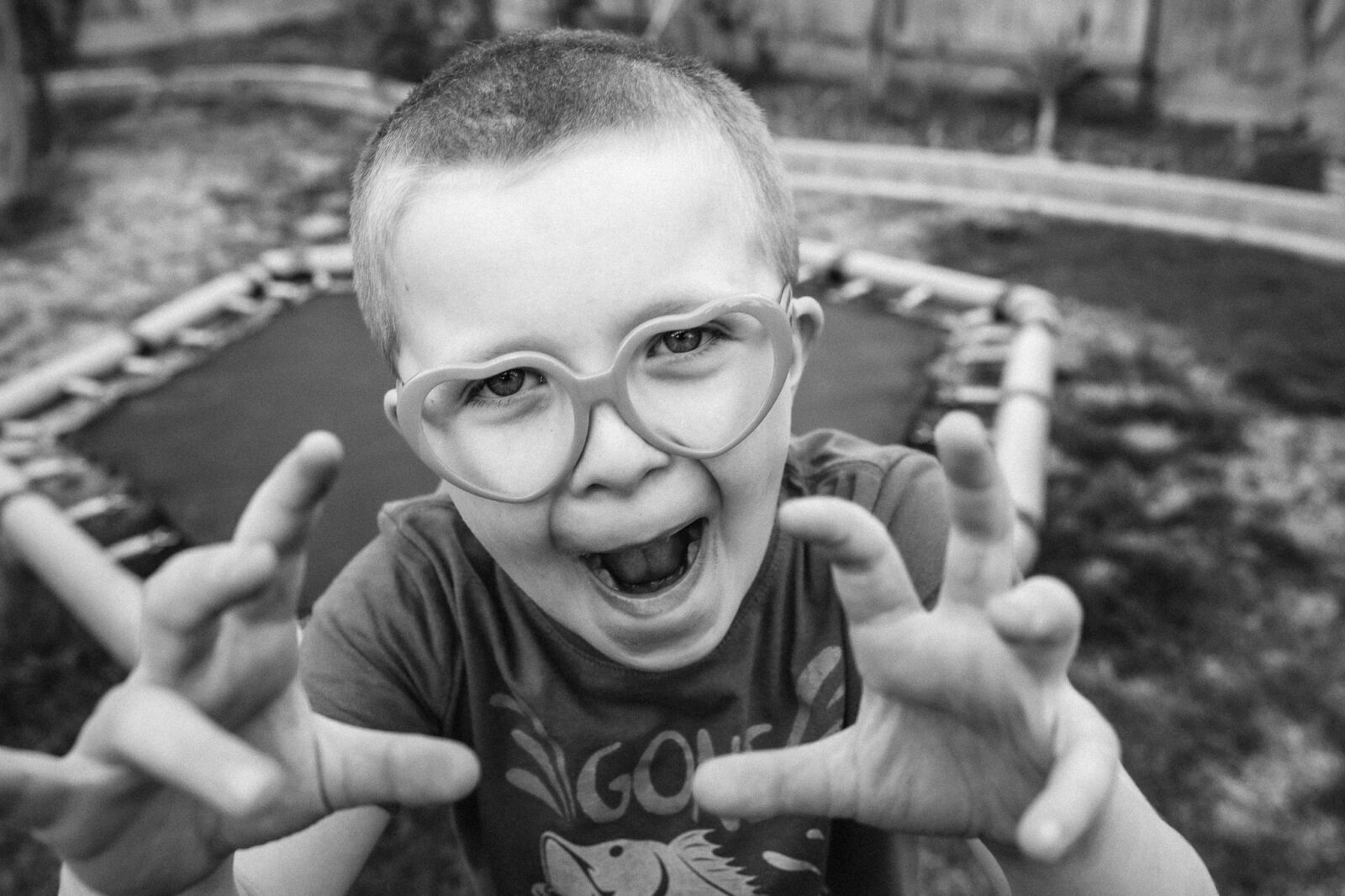 A little boy with heart-shaped glasses on reaches for the camera, making a silly face and hiding his teeth.